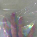Mão posicionada atrás de uma folha de foil furta-cor transparente, mostrando a translucidez do material e seus reflexos cintilantes com aparência de mancha de óleo